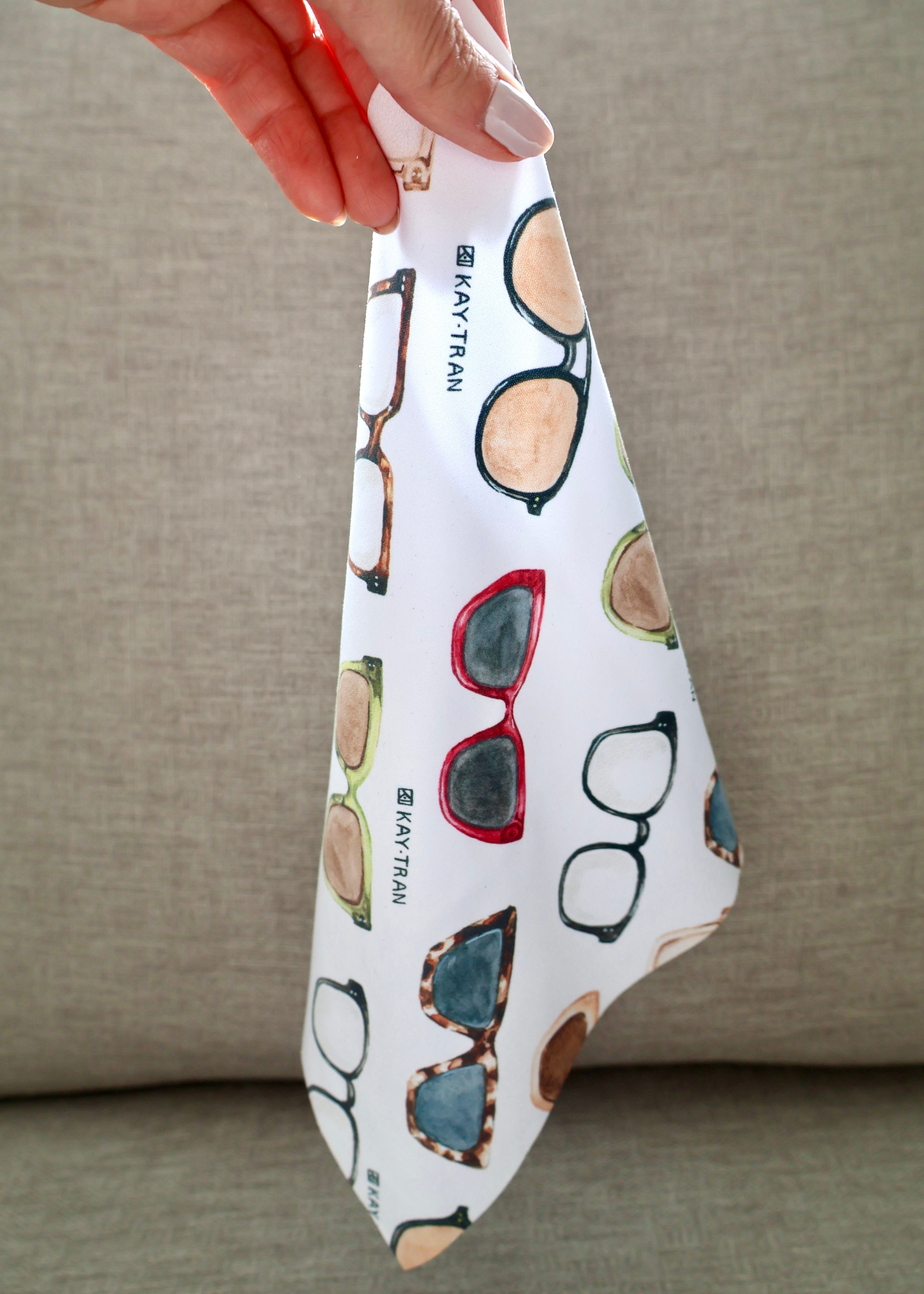 A hand holds the KayTran x Ton Ton Art Tune Up Kit cloth with colorful frames by KayTran Eyewear against a gray background.
