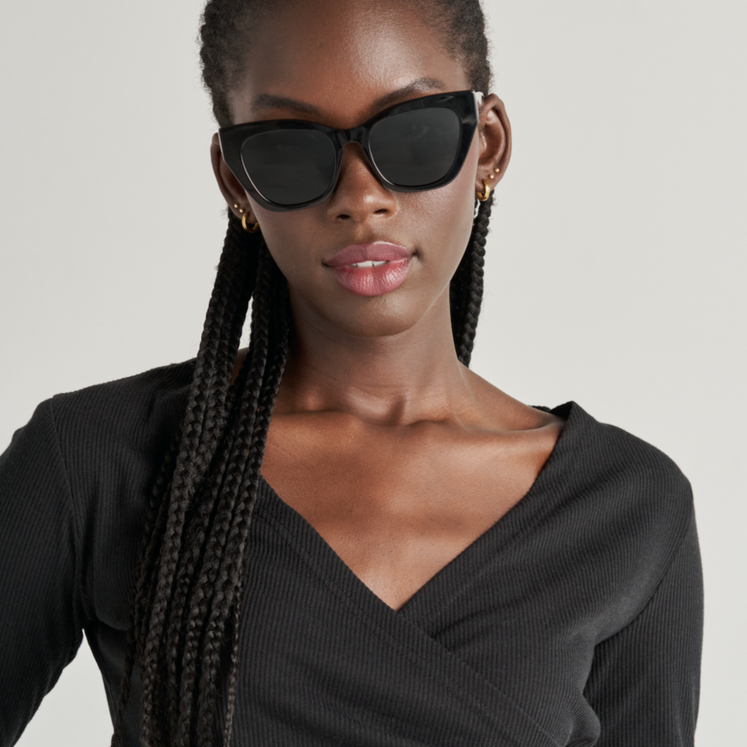 Woman with long braids wears KayTran Eyewear Harper - Polished Black sunglasses and a black V-neck top.