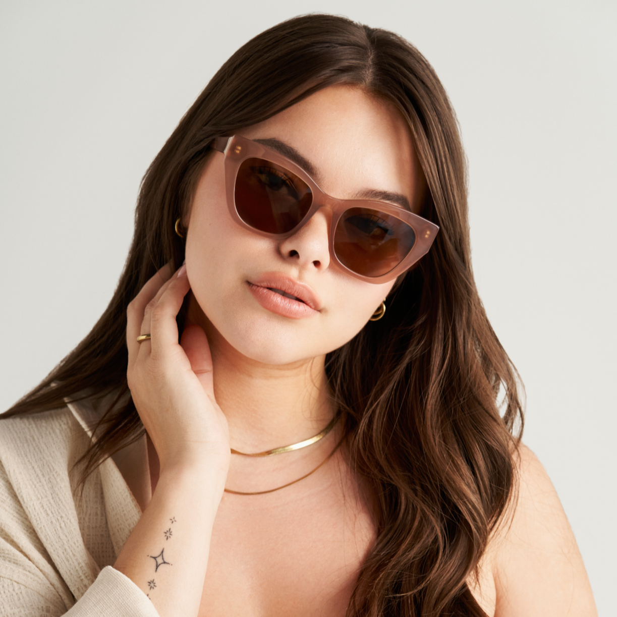 Woman with long brown hair wears KayTran Eyewear Harper - Almond cat-eye sunglasses and a beige top, touching her neck.