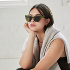 Woman wearing KayTran Eyewears Harper - Olive sunglasses and a beige sweater, sitting indoors with her hand on her chin.