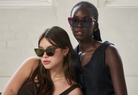 Two women in KayTran Eyewears Harper - Berry sunglasses pose together in front of a white brick wall.