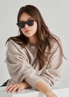 Woman in beige sweater wears KayTran Eyewears Frankie - Chocolate Chip sunglasses at a white table, plain background.