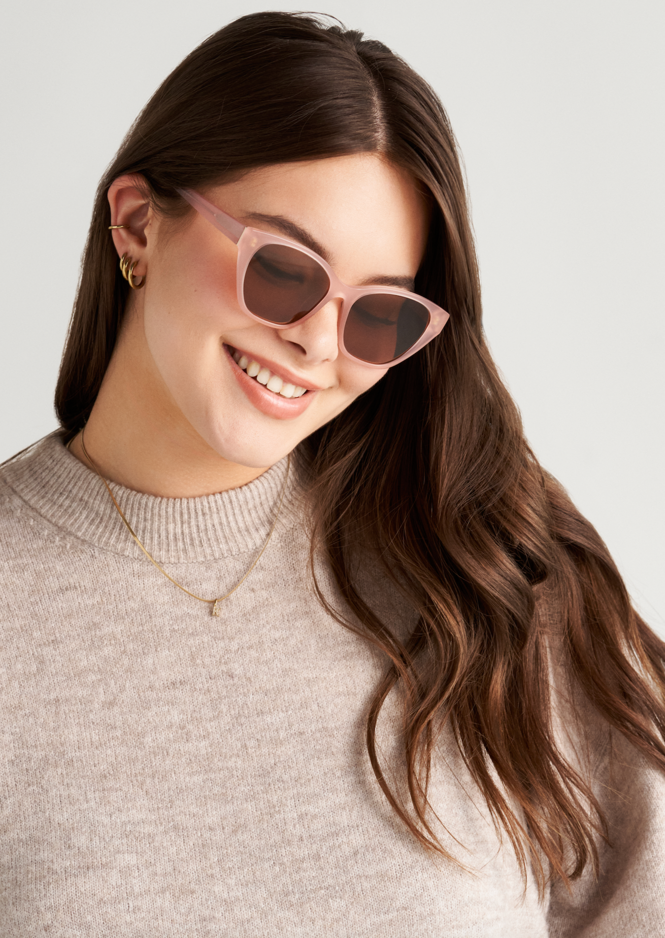 Woman with long brown hair smiles in KayTran Eyewears Taylor - Milky Blush cat-eye sunglasses and gold earrings.