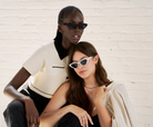 Two women in Naomi - Polished Black by KayTran Eyewear pose in neutral outfits with black accents by a white brick wall.