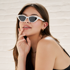 Woman wearing KayTran Eyewears Naomi - Birch cat-eye sunglasses, resting her chin on her hand by a white brick wall.