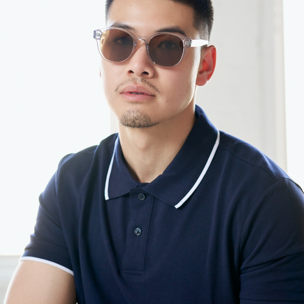 A man wears KayTran Eyewear’s Reese - Fog sunglasses and a navy polo, sitting by a bright window, facing the camera.