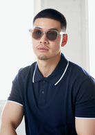 A man wears KayTran Eyewear’s Reese - Fog sunglasses and a navy polo, sitting by a bright window, facing the camera.