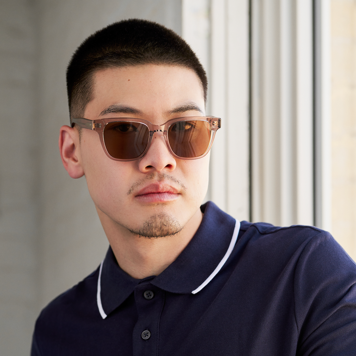 A man in a navy polo and KayTran Eyewear’s Charlie - Stone Taupe sunglasses stands by a window, looking aside.