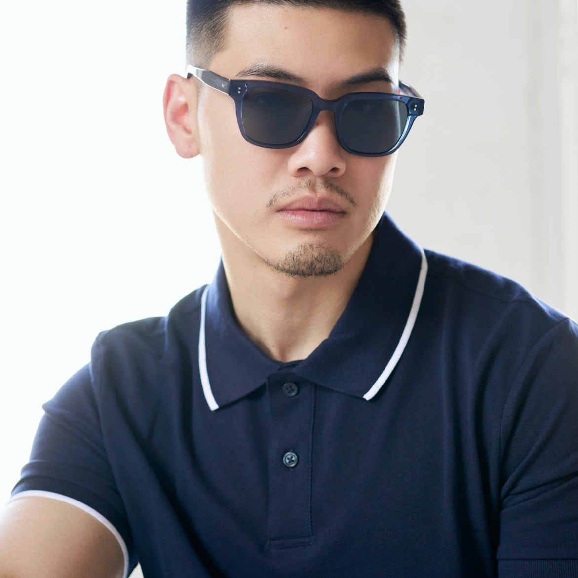 A man wears KayTran Eyewears Charlie - Dark Denim sunglasses with a navy polo against a bright, neutral background.
