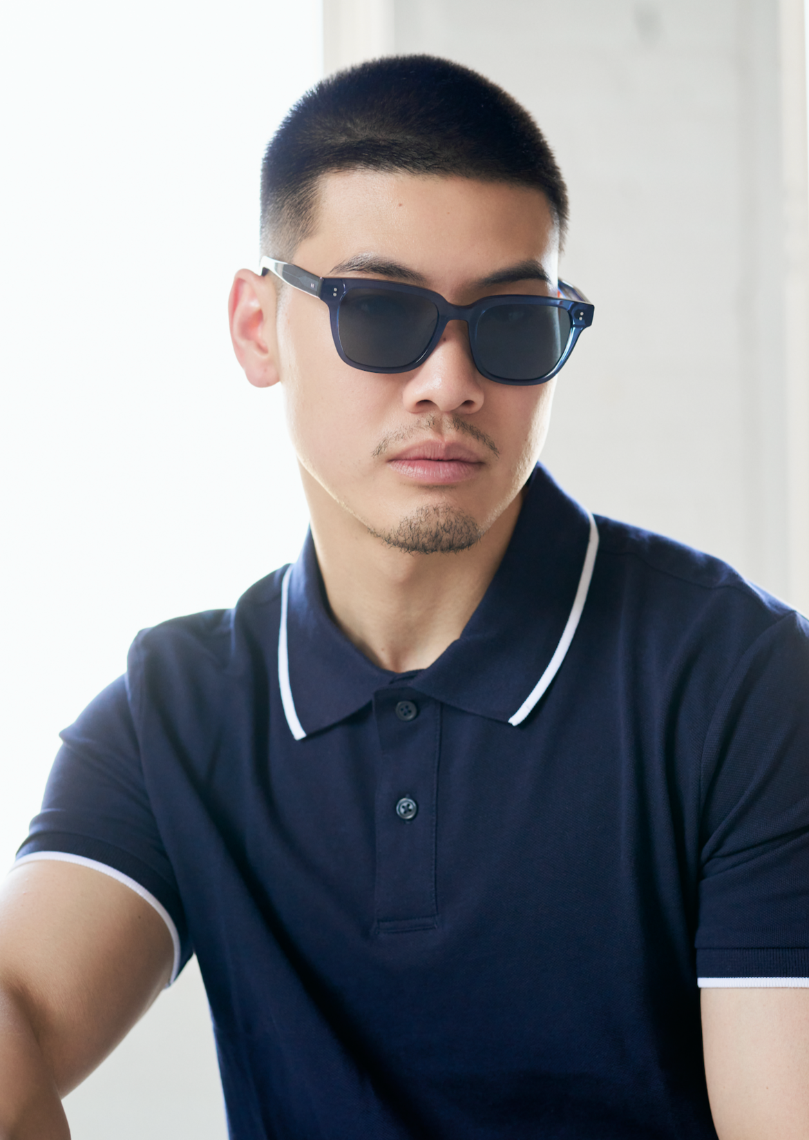 A man wears KayTran Eyewears Charlie - Dark Denim sunglasses with a navy polo against a bright, neutral background.