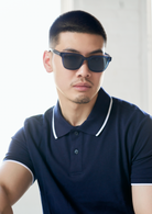 A man wears KayTran Eyewears Charlie - Dark Denim sunglasses with a navy polo against a bright, neutral background.