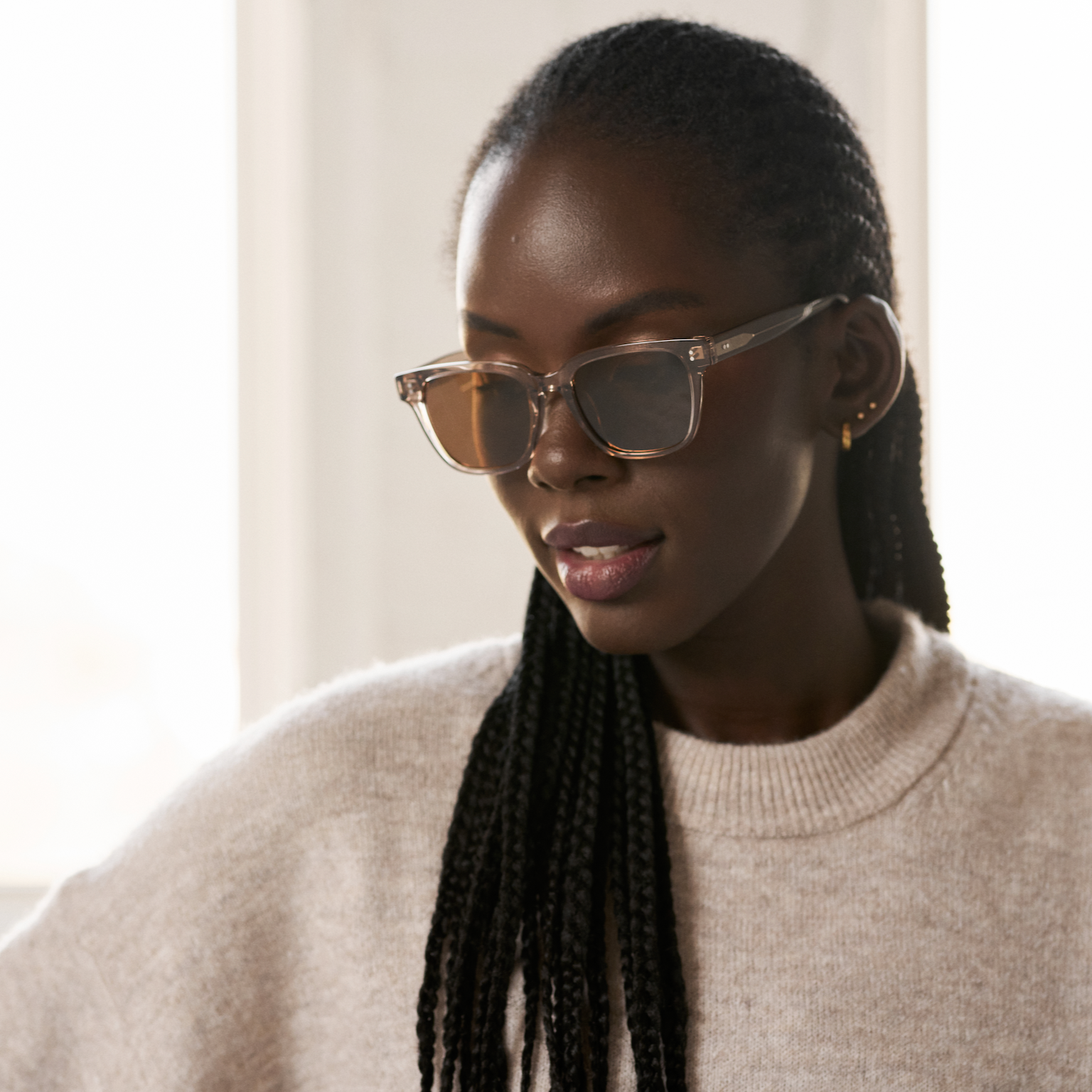 A woman with braided hair wears KayTran Eyewears Charlie - Stone Taupe sunglasses and a beige sweater, looking down.