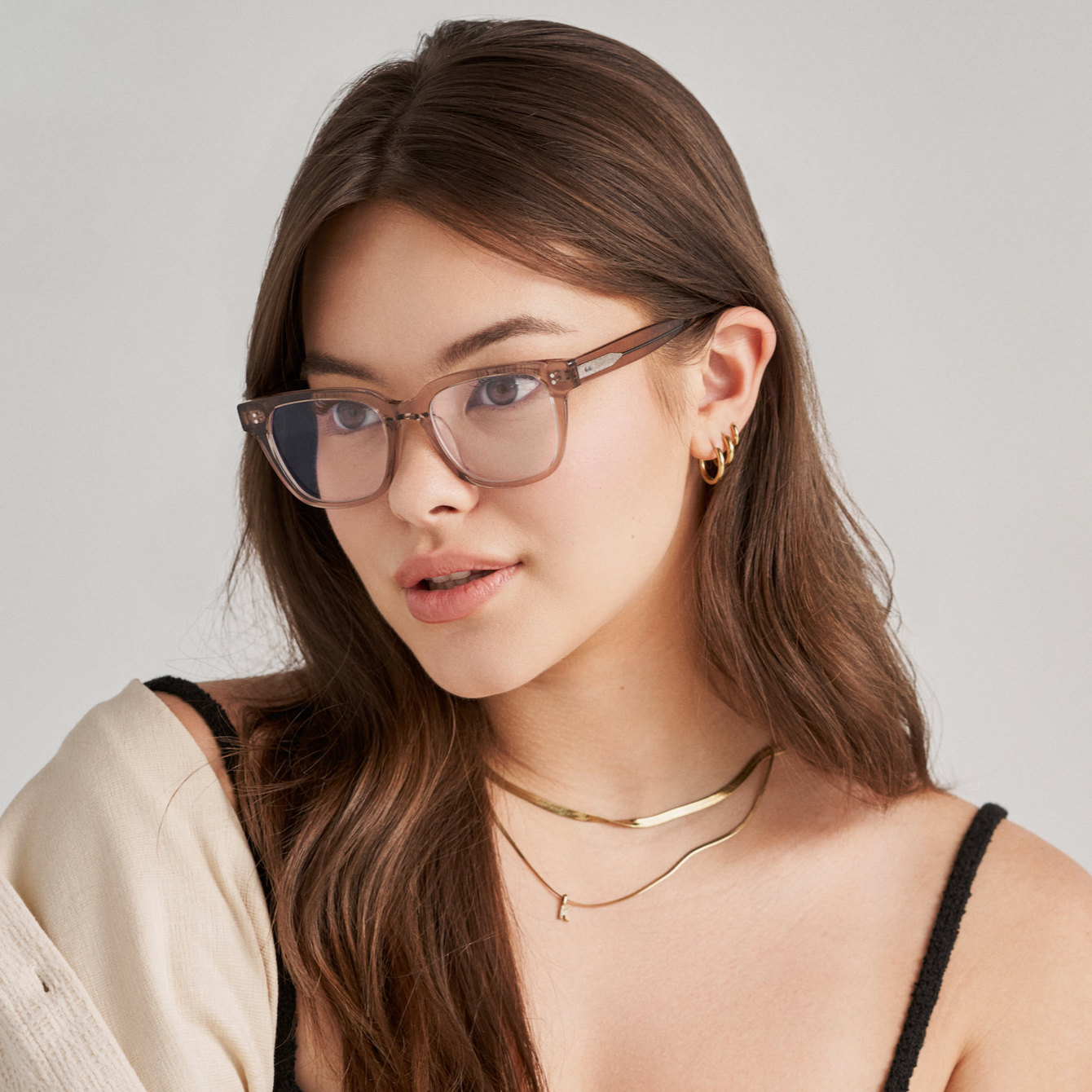 A woman with long brown hair wears KayTran Eyewear’s Charlie Optical - Stone Taupe and gold jewelry, gazing to the side.