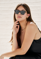 Woman in a black dress wears KayTran Eyewear’s Frankie - Chocolate Chip sunglasses, sitting by a white brick wall.