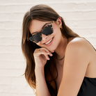 Woman with long brown hair wears KayTran Eyewears Frankie - Chocolate Chip sunglasses and a black top, smiling.