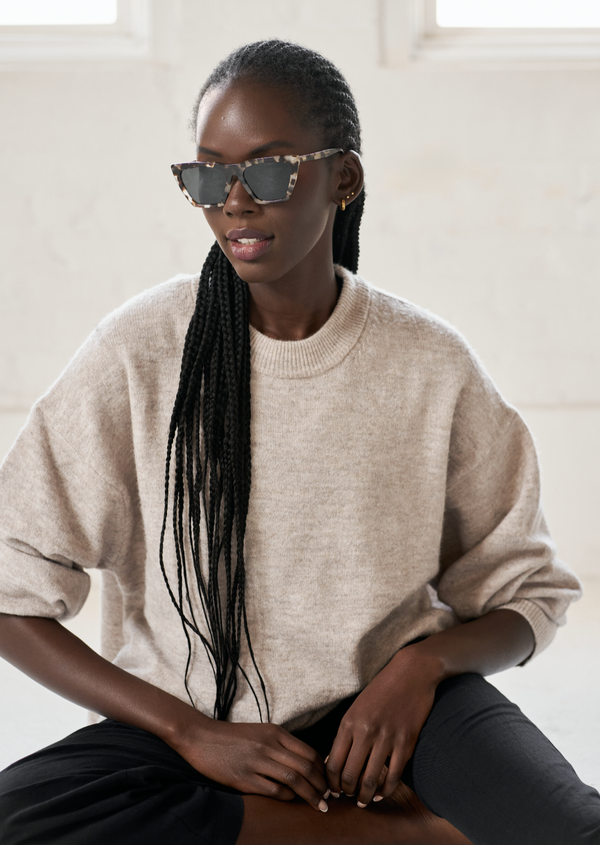 Woman with long braids wears KayTran Eyewear’s Frankie - Chocolate Chip sunglasses, beige sweater, and black pants indoors.