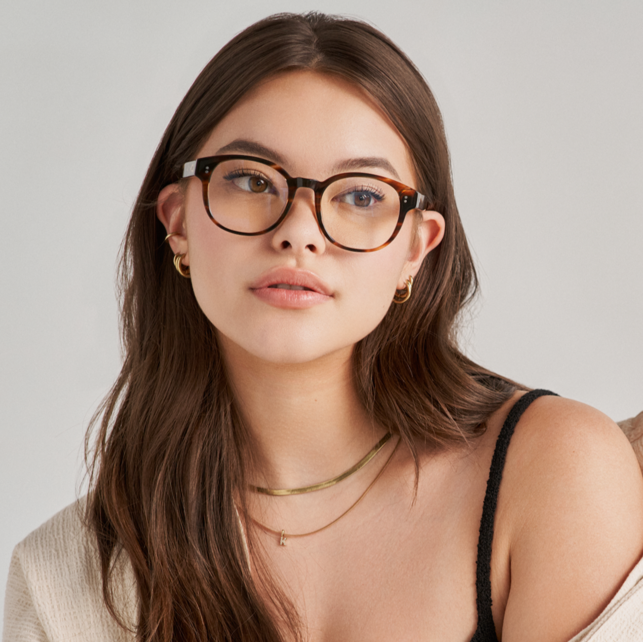 Young woman with long brown hair wears KayTran Eyewears Reese Optical - Amber Honey glasses, gold jewelry, and a beige cardigan.