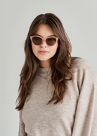Woman with long brown hair wears KayTran Eyewears Taylor - Milky Blush sunglasses, beige sweater, grey background.