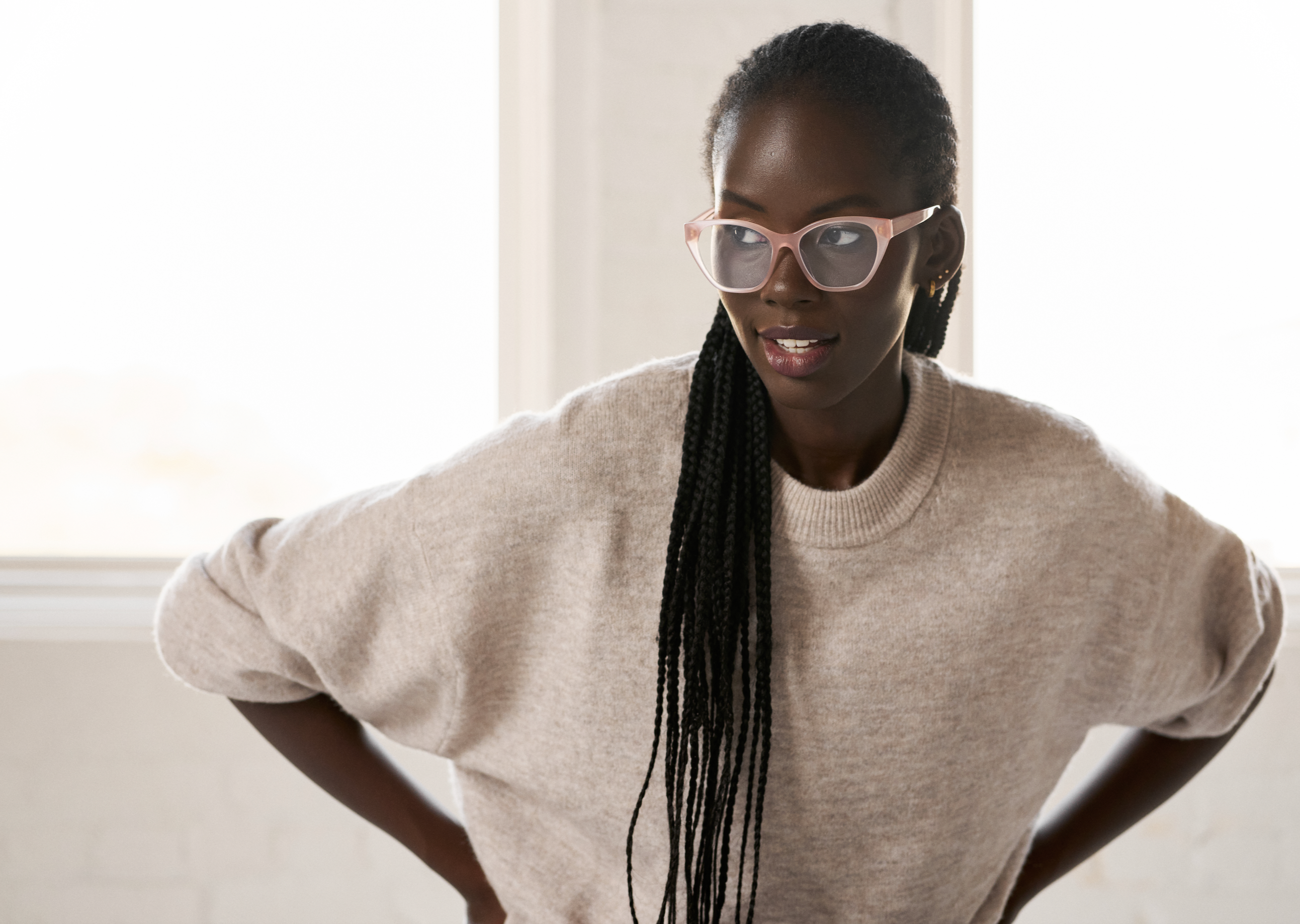 Woman wears KayTran Eyewears Taylor Optical - Milky Blush glasses and a beige sweater, standing hands on hips indoors.