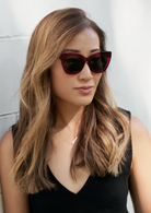 Woman with long, light brown hair wears KayTran Eyewear Arlo - Berry sunglasses and a black top against a white wall.