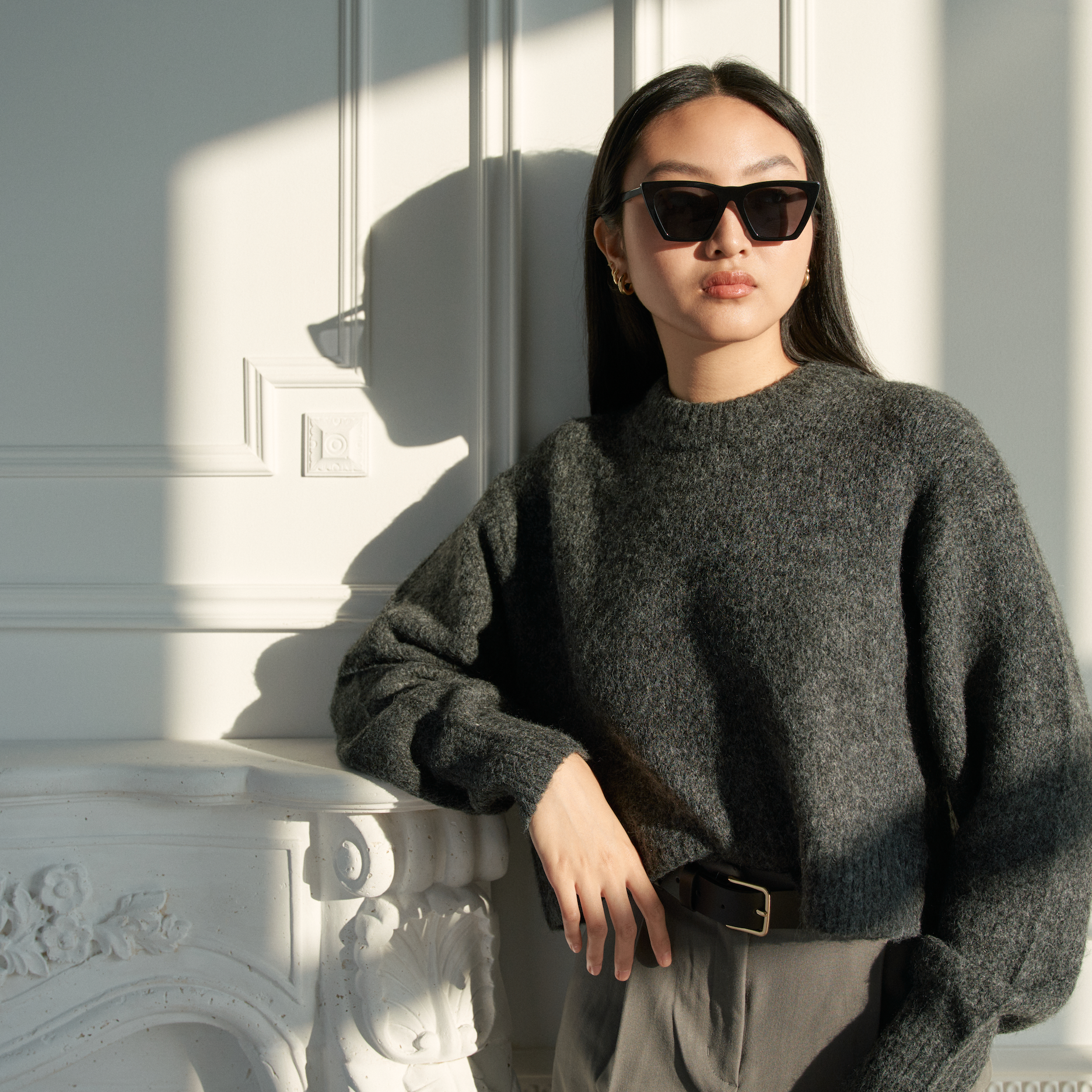 Woman wears KayTran Eyewears Frankie - Polished Black sunglasses, leaning by a white mantel in a sunlit room.