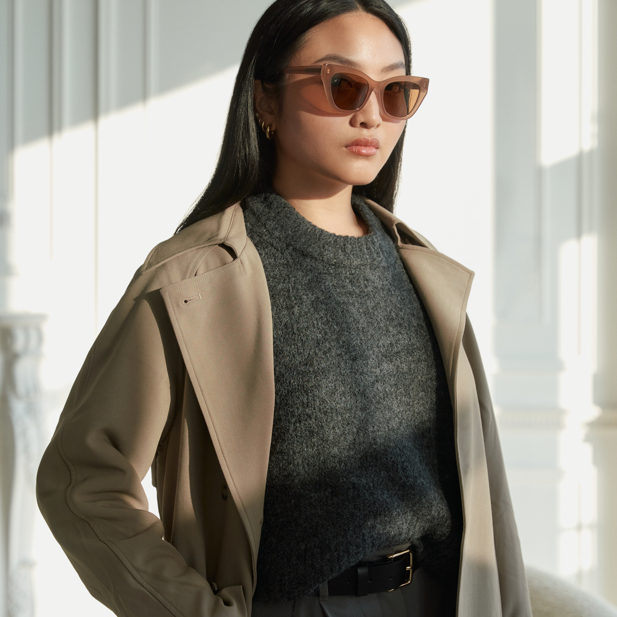 A woman wears KayTran Eyewears Harper - Almond sunglasses, a beige coat, and a dark sweater indoors with sunlight.