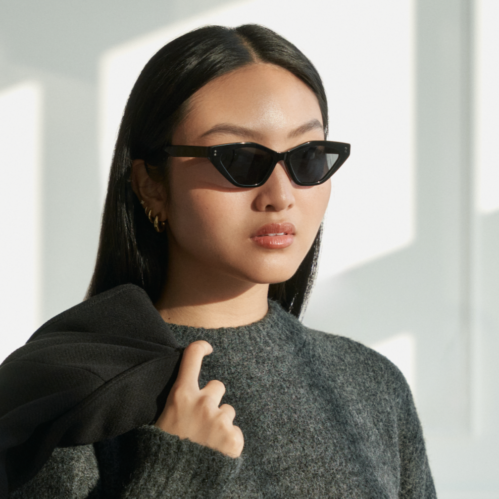 Woman wears KayTran Eyewears Naomi - Polished Black cat-eye sunglasses and a grey sweater, indoors with black coat.