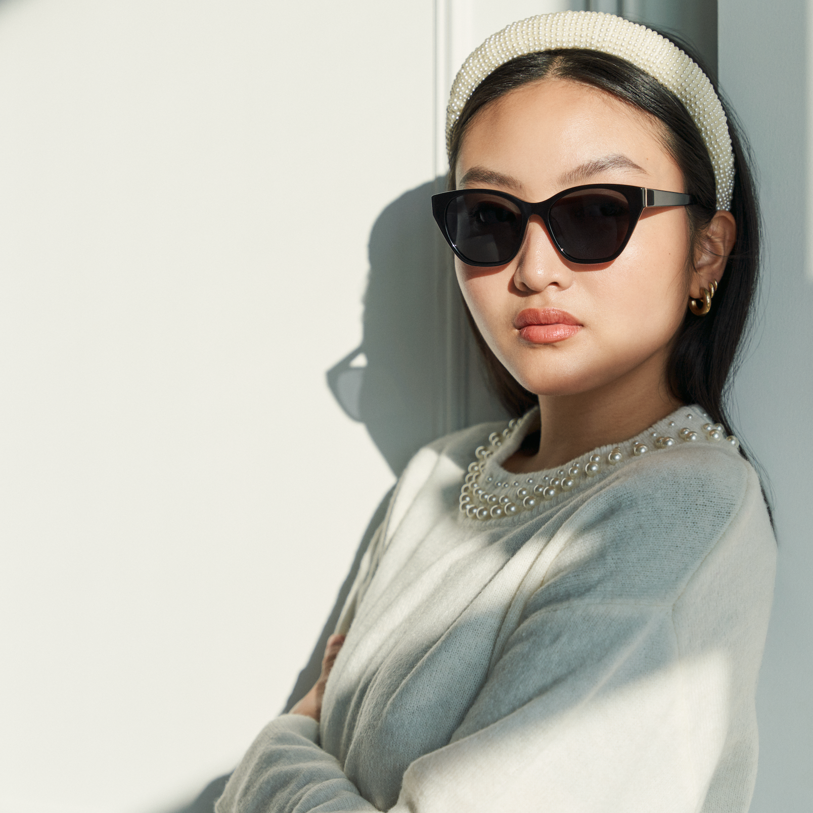A woman in KayTran Eyewears Taylor - Polished Black sunglasses stands by a white wall in natural light.