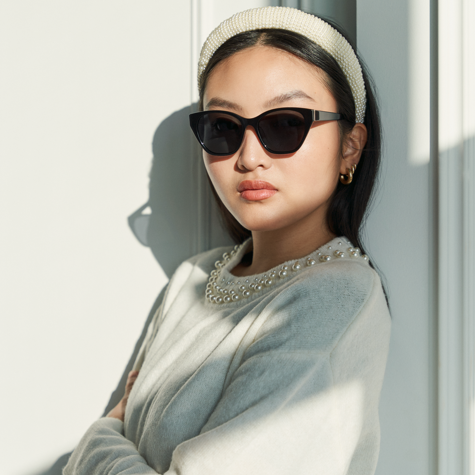 A woman in KayTran Eyewears Taylor - Polished Black sunglasses stands by a white wall in natural light.