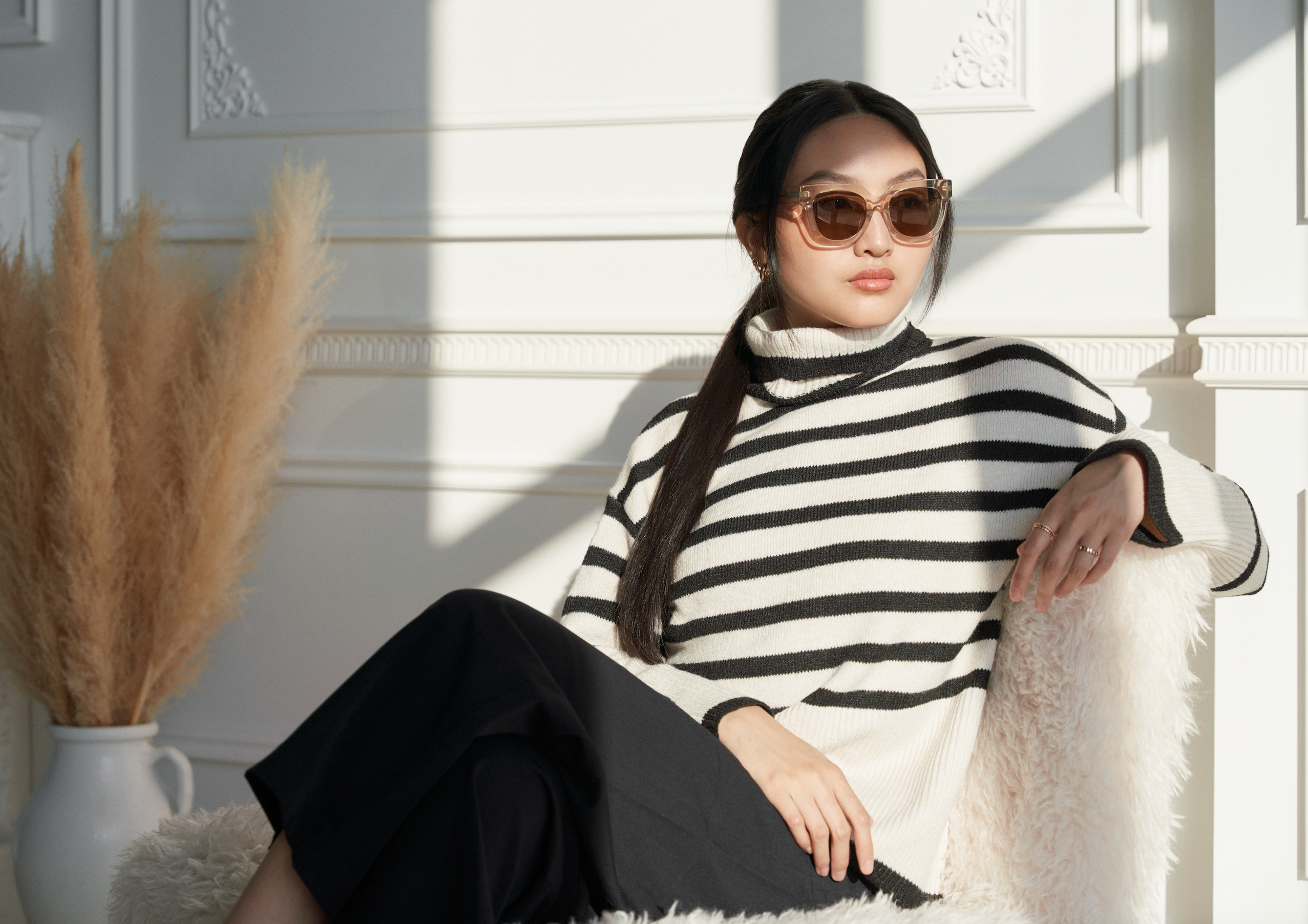 A woman in KayTran Eyewears Rowan – Sand sunglasses sits on a furry chair beside pampas grass.
