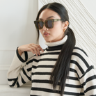 Woman wears KayTran Eyewear’s Rowan - Olive sunglasses, paired with a black-and-white striped turtleneck indoors.