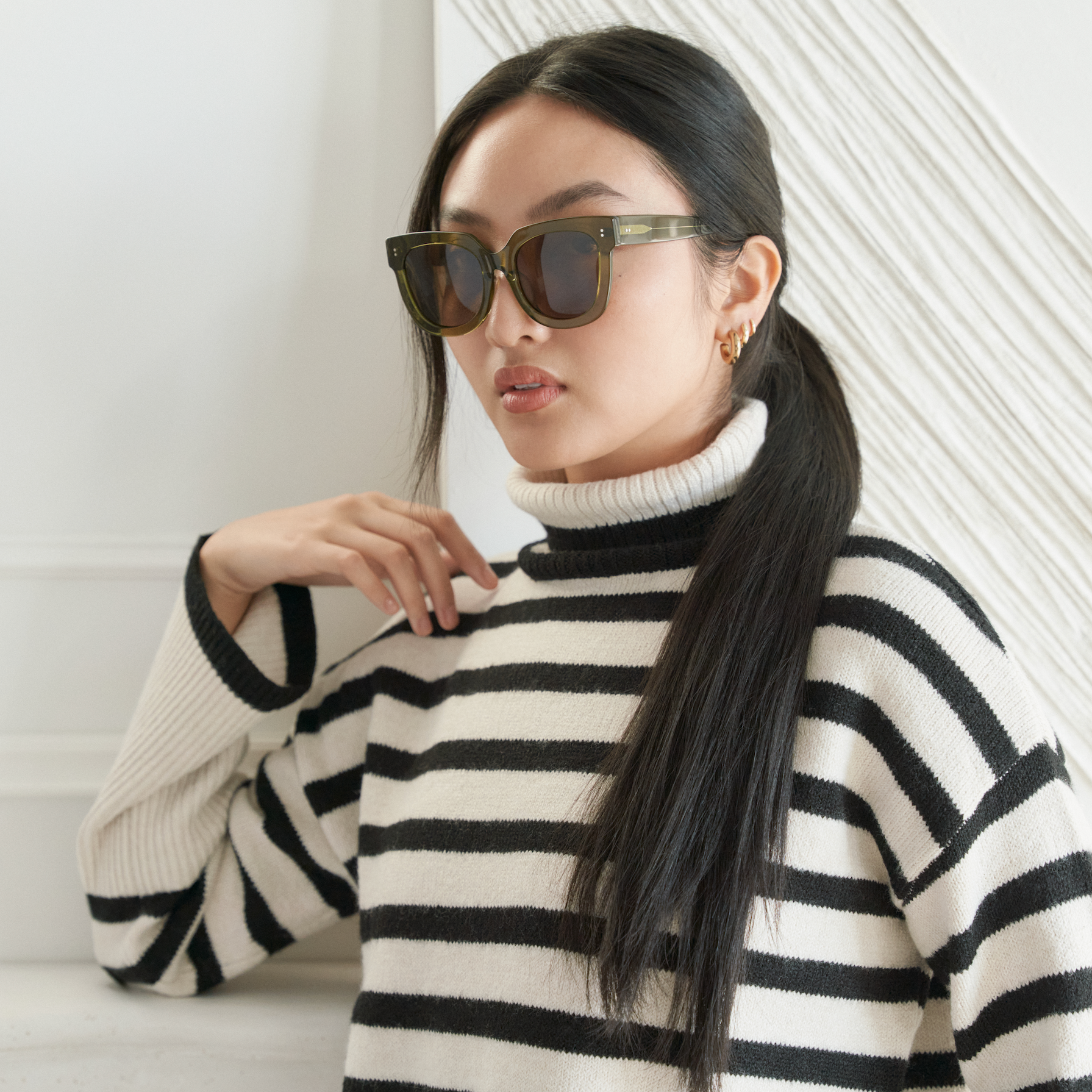 Woman wears KayTran Eyewear’s Rowan - Olive sunglasses, paired with a black-and-white striped turtleneck indoors.