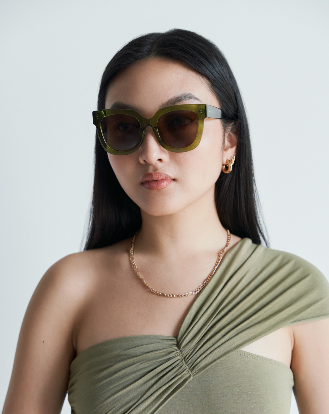 Woman wearing KayTran Eyewear Rowan - Olive sunglasses, gold jewelry, and an asymmetrical olive top against a plain background.