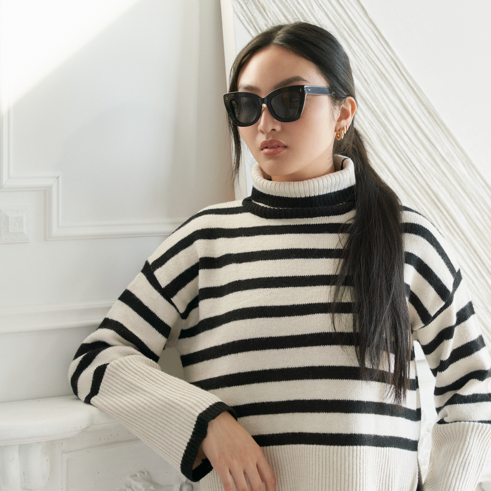 Woman wears KayTran Eyewear Arlo - Polished Black sunglasses and a striped turtleneck, standing indoors by a white wall.