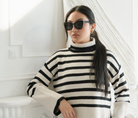 Woman wears KayTran Eyewear Arlo - Polished Black sunglasses and a striped turtleneck, standing indoors by a white wall.