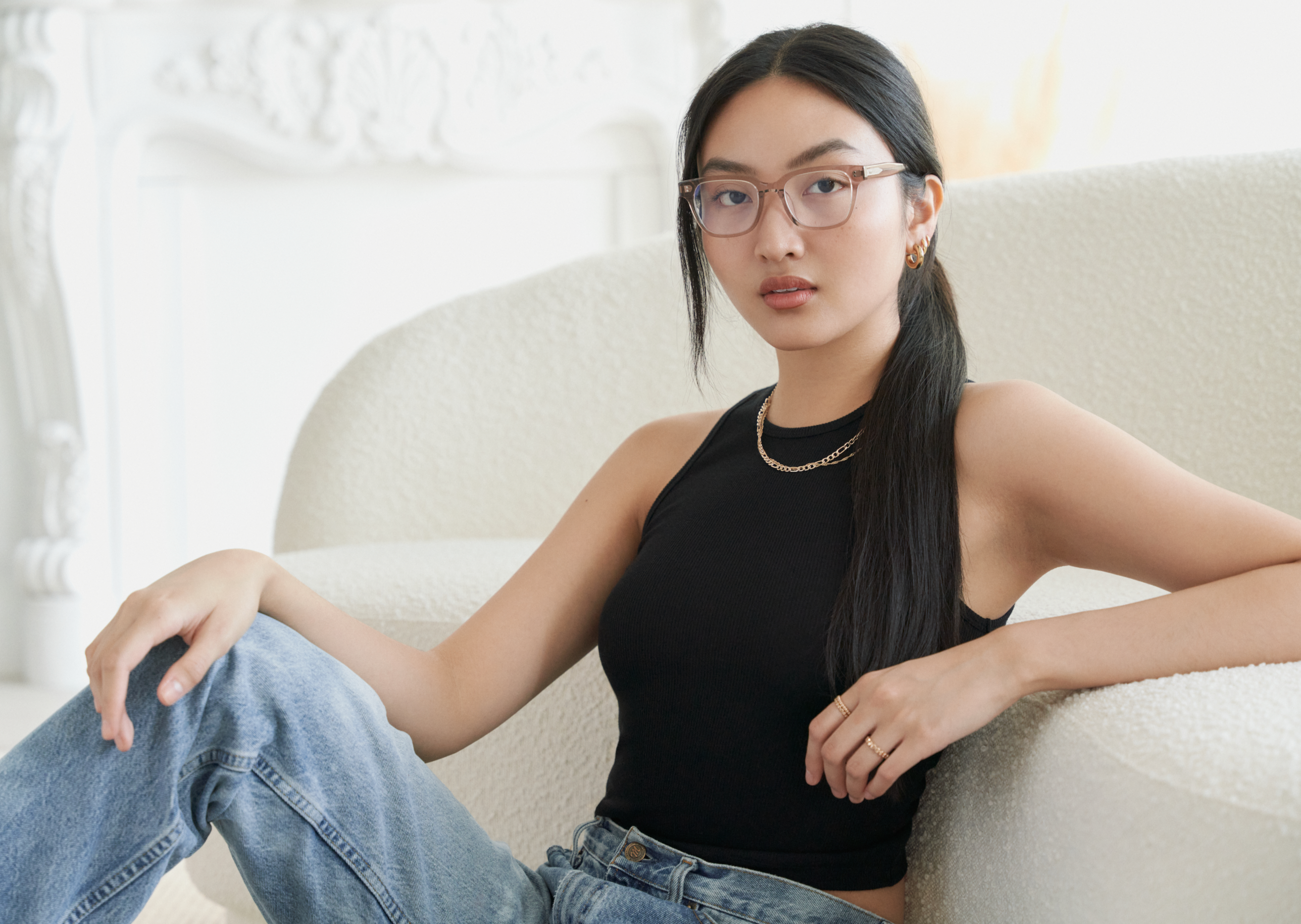 A woman in KayTran Eyewear’s Charlie Optical - Stone Taupe glasses sits on a light couch in a bright room.