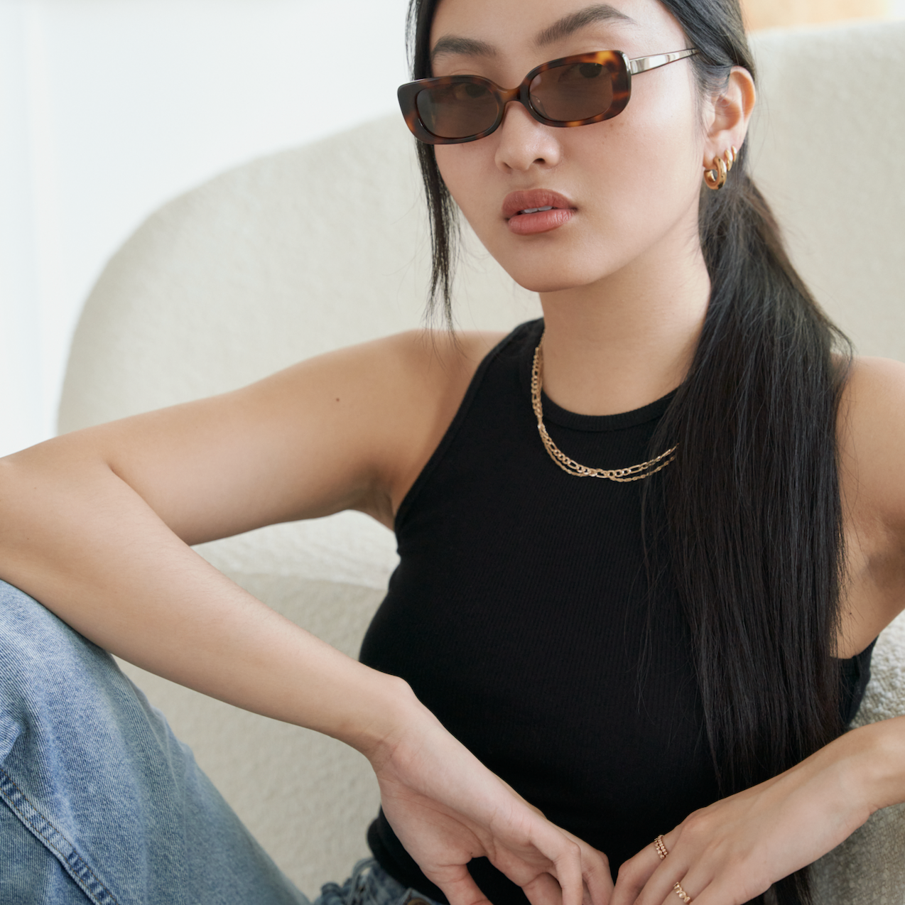 Woman wearing KayTran Eyewear Billie - Lava Tortoise sunglasses, black tank top, and jeans sits on a chair.