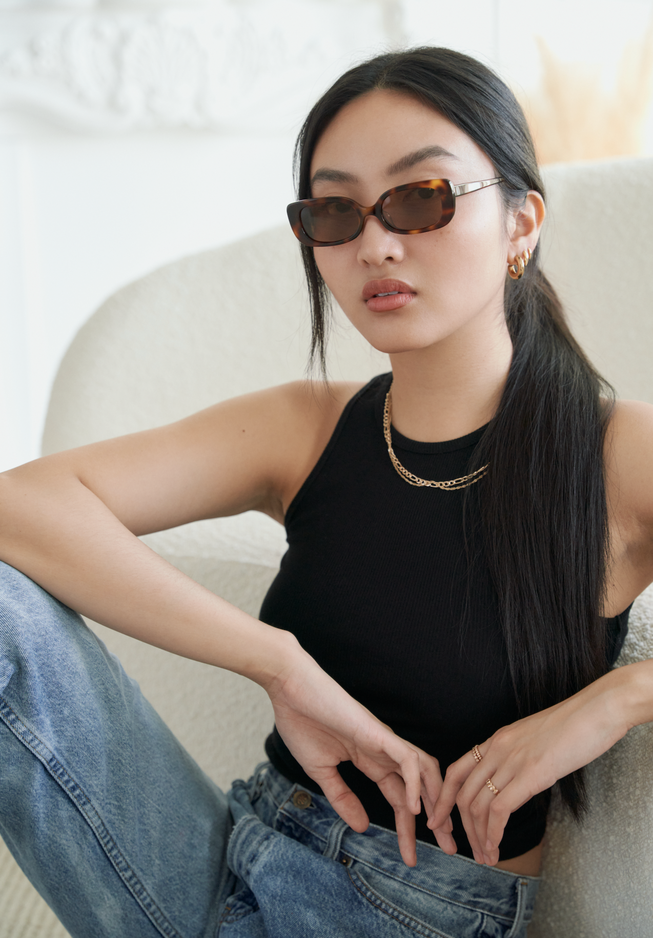 Woman wearing KayTran Eyewear Billie - Lava Tortoise sunglasses, black tank top, and jeans sits on a chair.