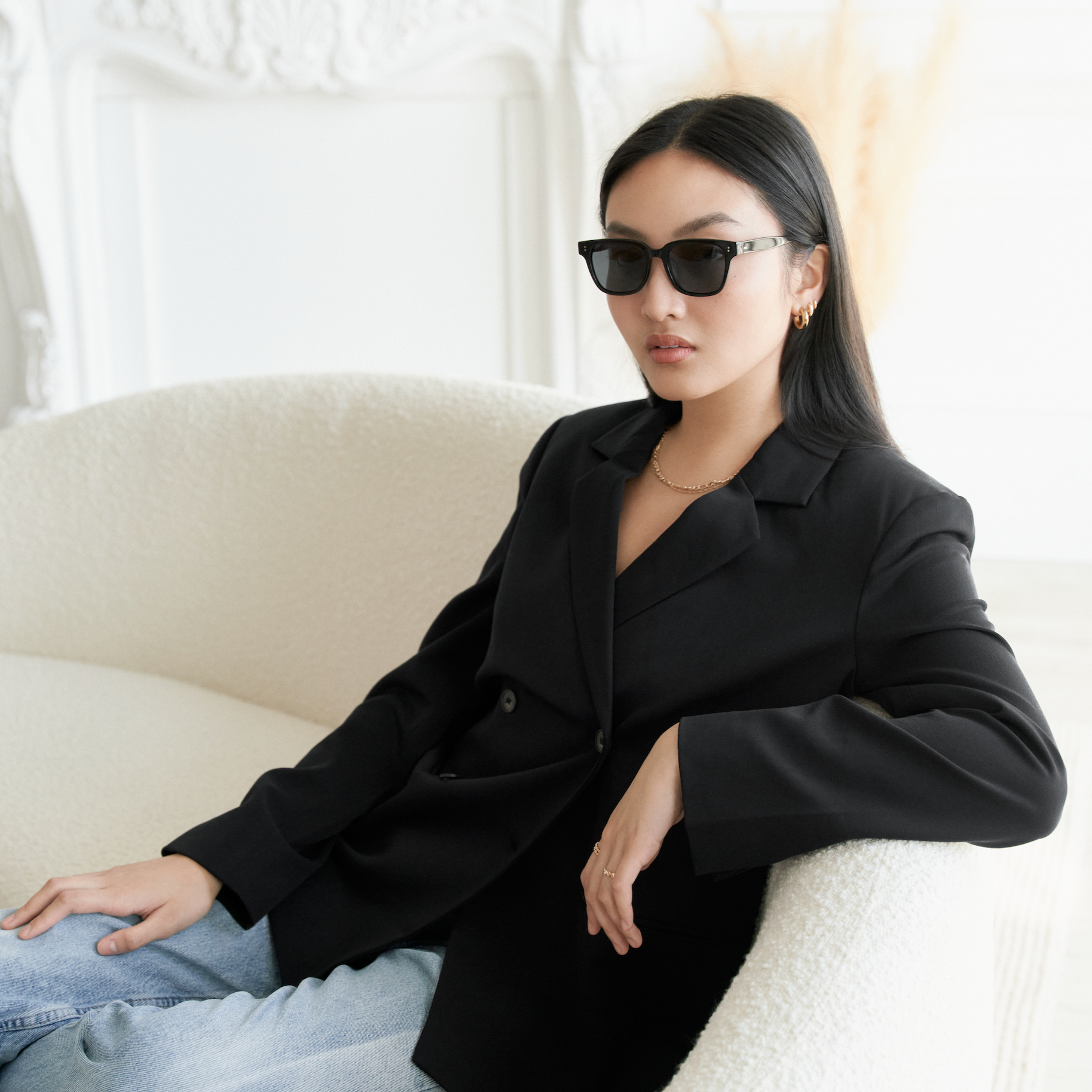 Woman wears KayTran Eyewear Charlie - Polished Black sunglasses, sitting on a white textured sofa in a bright room.