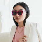 Woman in Arlo - Berry sunglasses by KayTran Eyewear, white blazer, pink sweater, gold chain, sitting indoors.