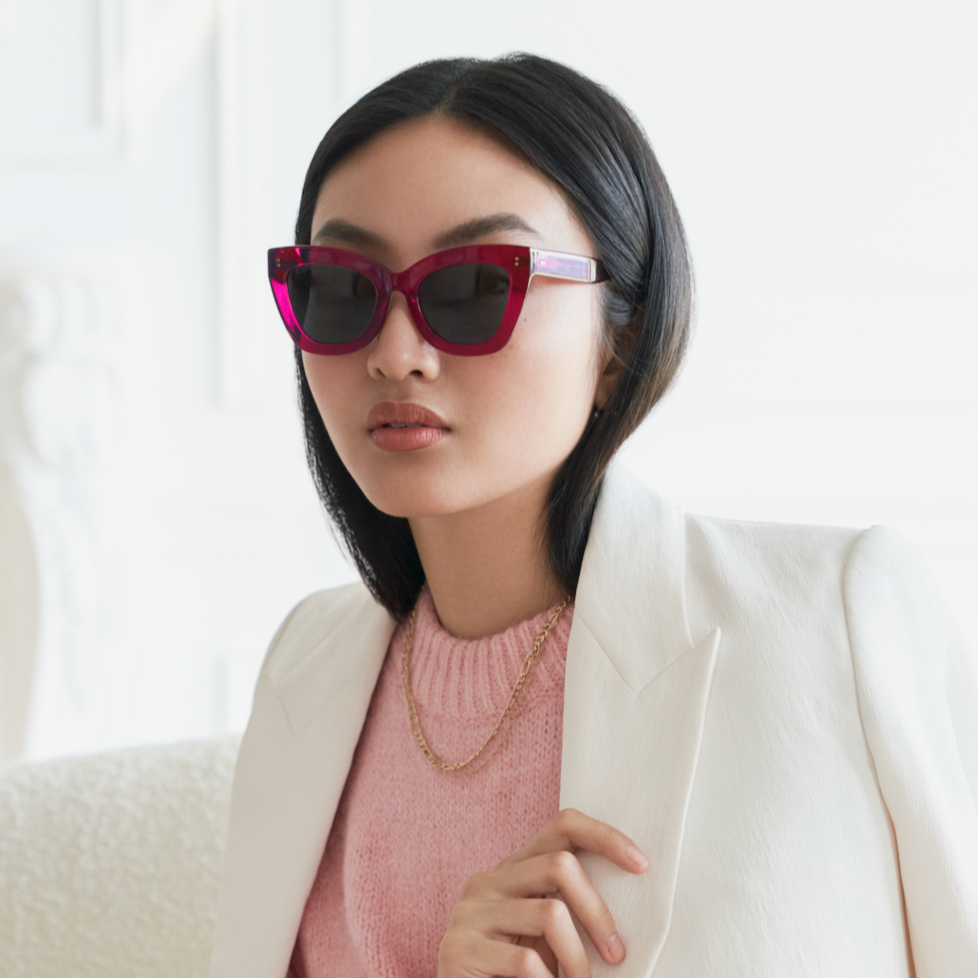 Woman in Arlo - Berry sunglasses by KayTran Eyewear, white blazer, pink sweater, gold chain, sitting indoors.