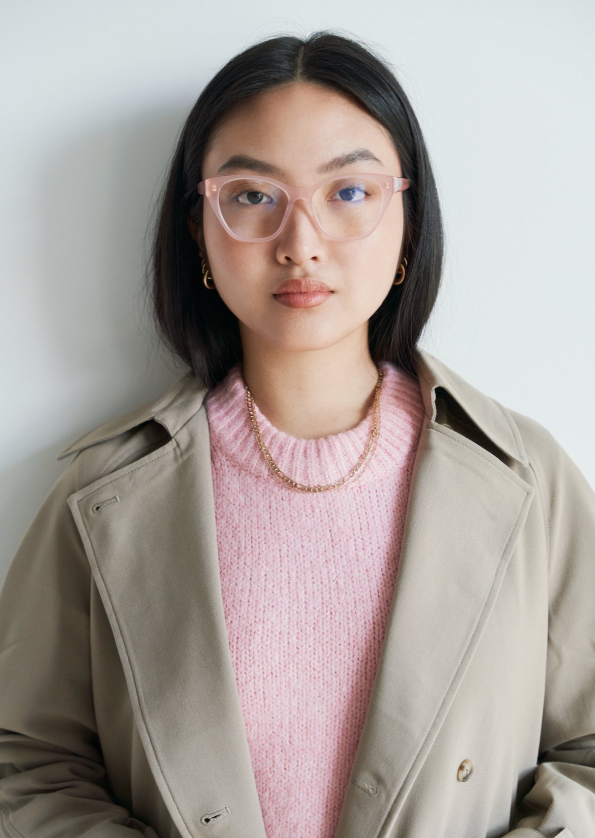 Woman with straight black hair wears KayTran Eyewear Taylor Optical - Milky Blush glasses, beige coat, and pink sweater.