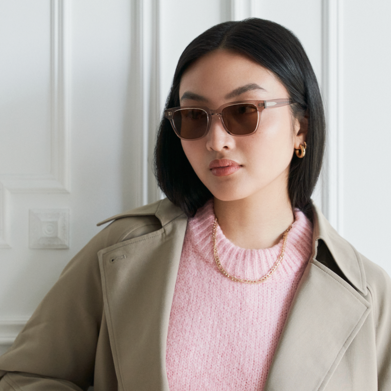 Woman with straight black hair wearing KayTran Eyewear Charlie - Stone Taupe sunglasses, pink sweater, and beige coat.