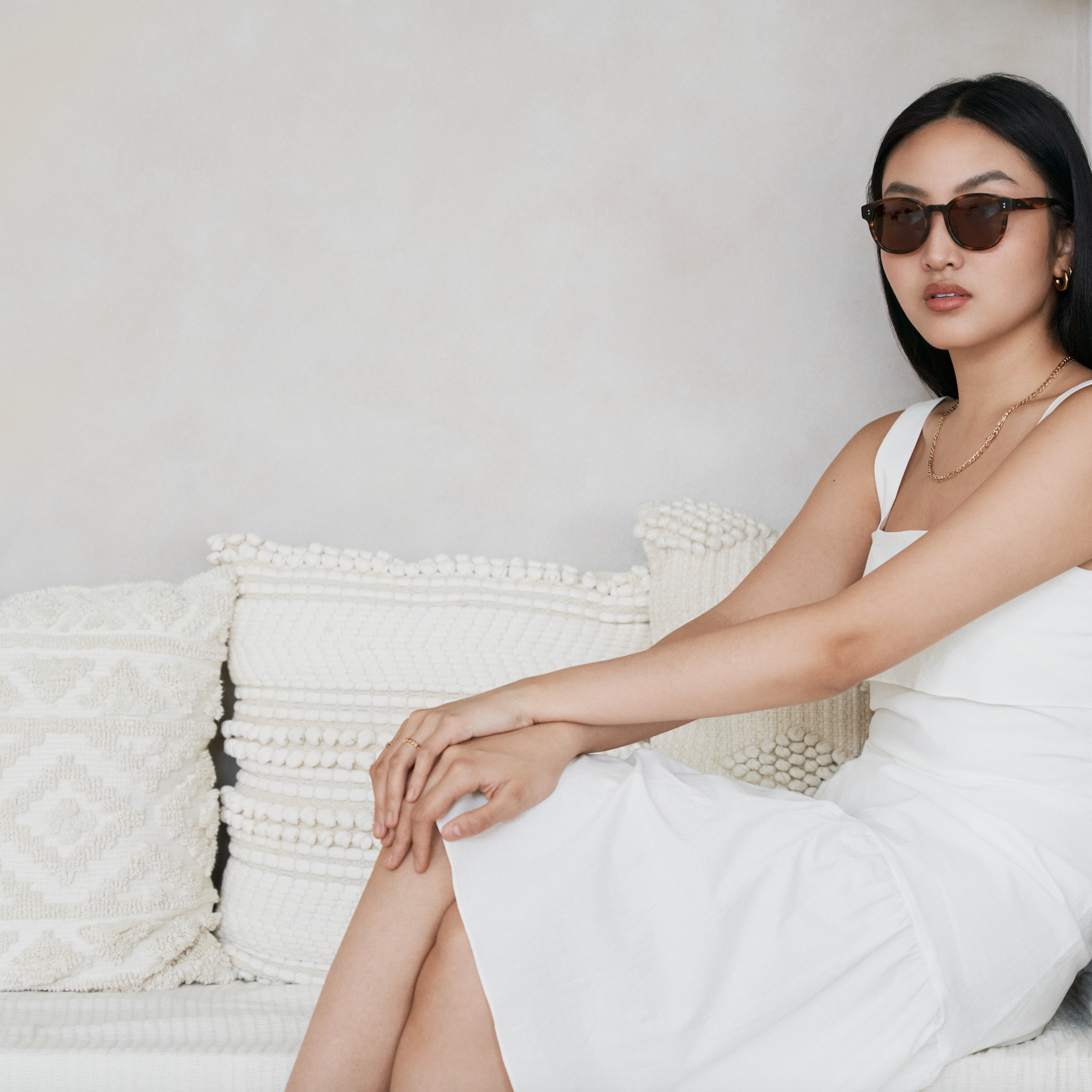 Woman in a white dress wears KayTran Eyewears Reese - Amber Honey sunglasses on a bench with textured pillows.