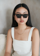 Woman with long dark hair wearing KayTran Eyewear Frankie - Chocolate Chip sunglasses, white sleeveless top, and gold jewelry.