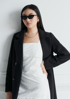 Woman in a black coat and white dress wears KayTran Eyewear Naomi - Polished Black sunglasses and gold jewelry.