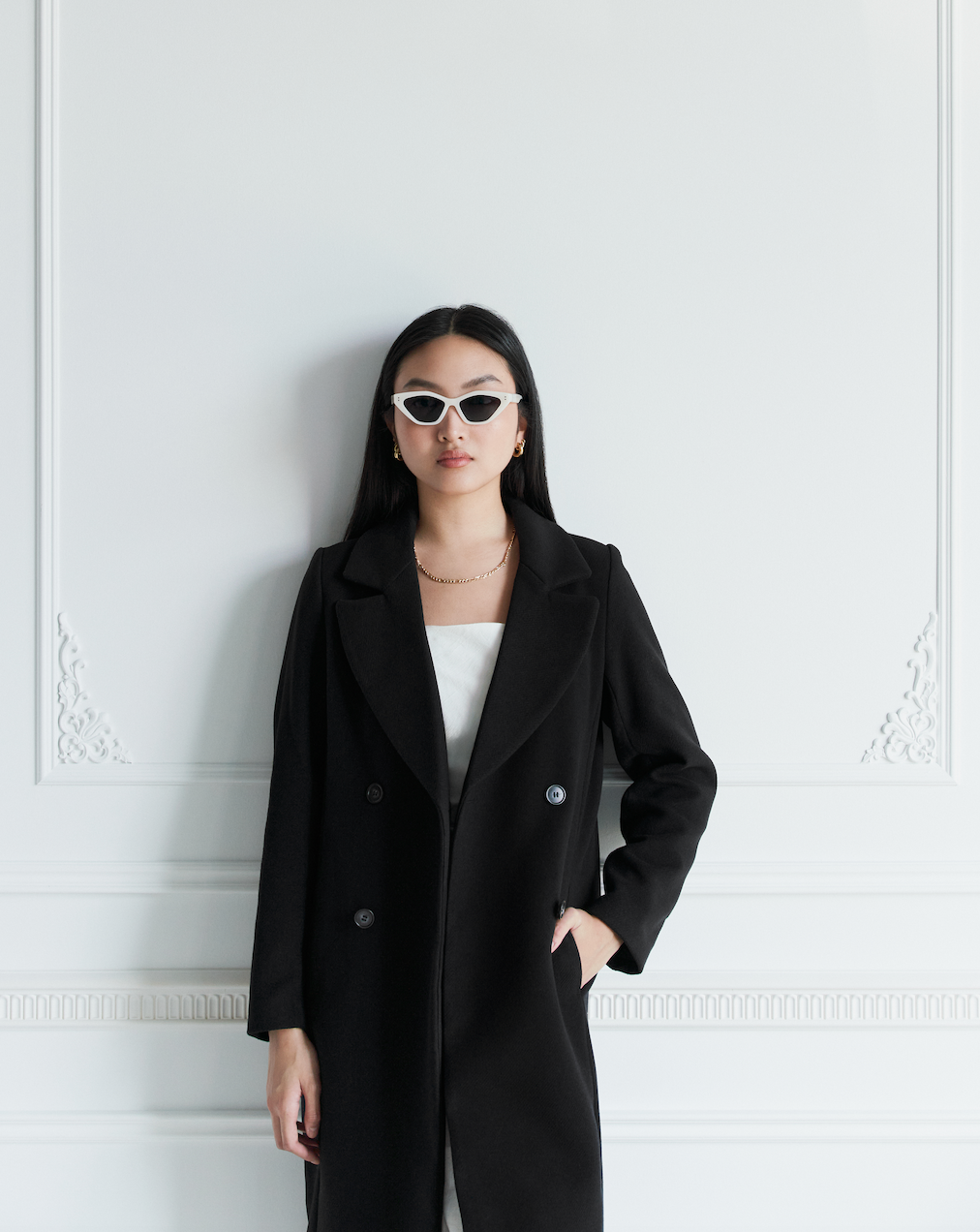 Woman wearing KayTran Eyewears Naomi - Birch sunglasses, standing against a white ornate wall, facing the camera.