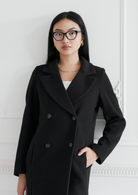 Woman in black double-breasted coat wears KayTran Eyewears Taylor Optical - Polished Black, standing by a white wall.