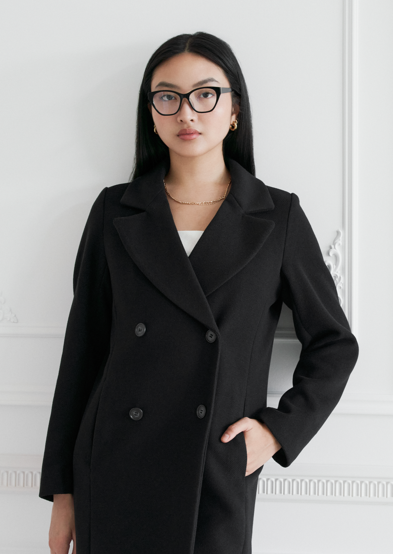 Woman in black double-breasted coat wears KayTran Eyewears Taylor Optical - Polished Black, standing by a white wall.