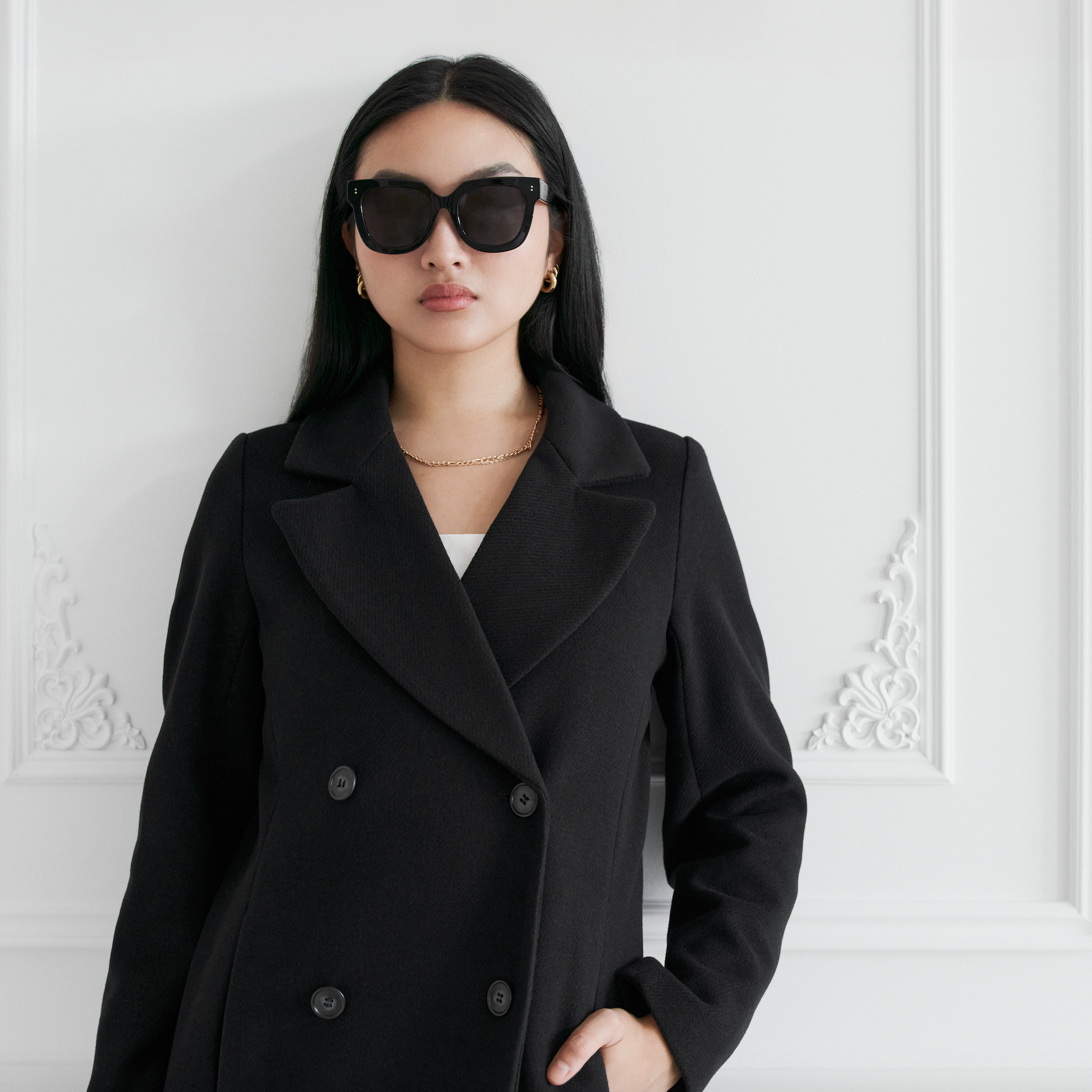 Woman wears KayTran Eyewears Rowan - Polished Black sunglasses and stands by an ornate white wall.